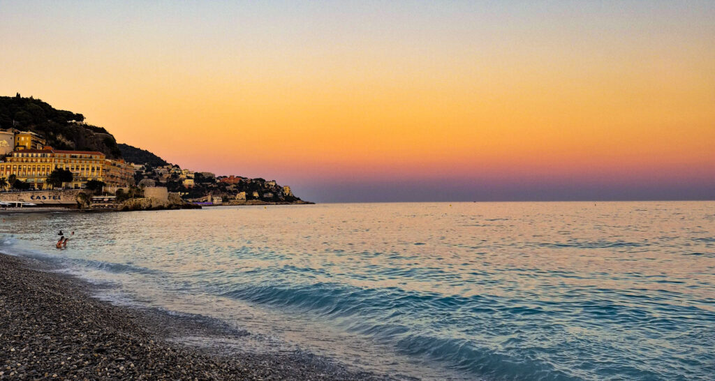 Pebble beach in Nice, France, at sunset, with waves rolling onto the shore and the historic Castel Plage in the background.