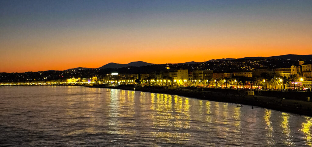 Nighttime view of the Baie des Anges in Nice, with city lights reflecting on the water and the Alps silhouetted against an orange horizon.