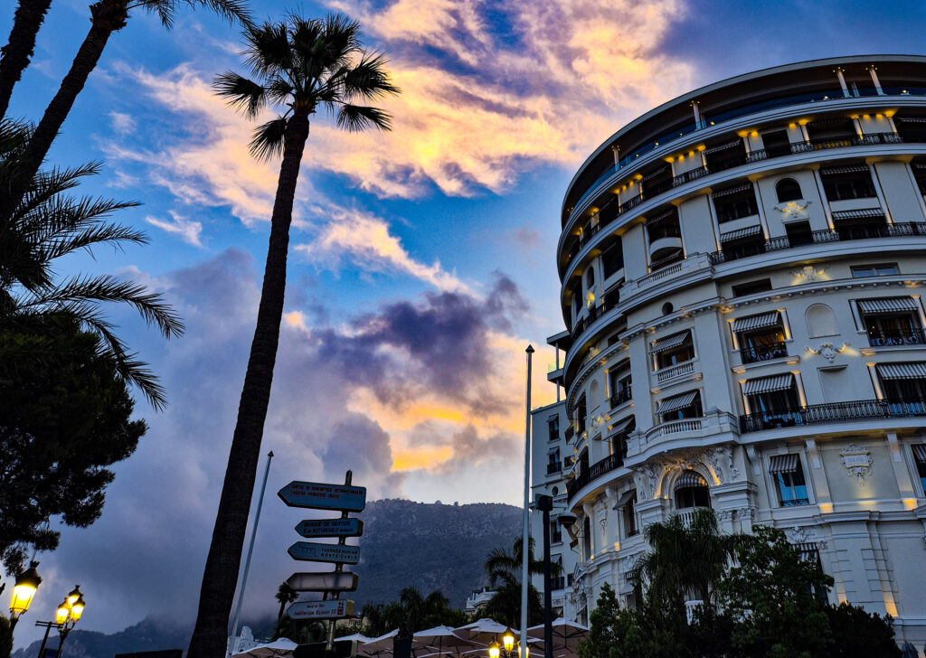 Monaco at sunset taken from behind the Hotel de Paris, feturing palm trees and a purplee-toned sunset sky.